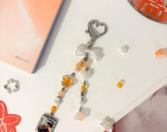 May include: A silver-toned keychain with a heart-shaped clasp and a white bow. The charm features a small framed photo, yellow star and flower-shaped beads, and dangling accents. The background includes a CD, a book, and scattered decorative elements.