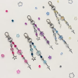 May include: Several beaded keychains with silver-tone hardware and charms. Each keychain features a different color scheme of beads, including pink, purple, blue, and yellow. Charms include stars, flowers, and a crescent moon. Scattered around are star and flower-shaped charms.