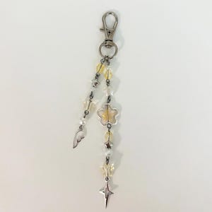 May include: A silver-toned keychain with a lobster clasp, adorned with yellow and white beads, star and flower charms, and a small wing. The keychain is designed with a celestial theme.