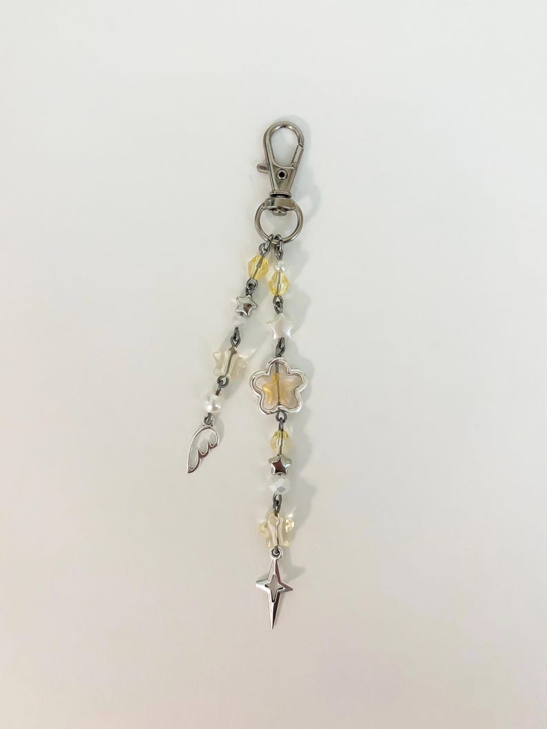 May include: A silver-toned keychain with a lobster clasp, adorned with yellow and white beads, star and flower charms, and a small wing. The keychain is designed with a celestial theme.