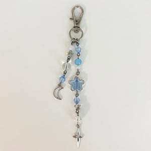 May include: A silver-toned keychain with a lobster clasp. The charm features a crescent moon, a blue flower, a star, and clear and blue beads. The keychain is designed with celestial elements.
