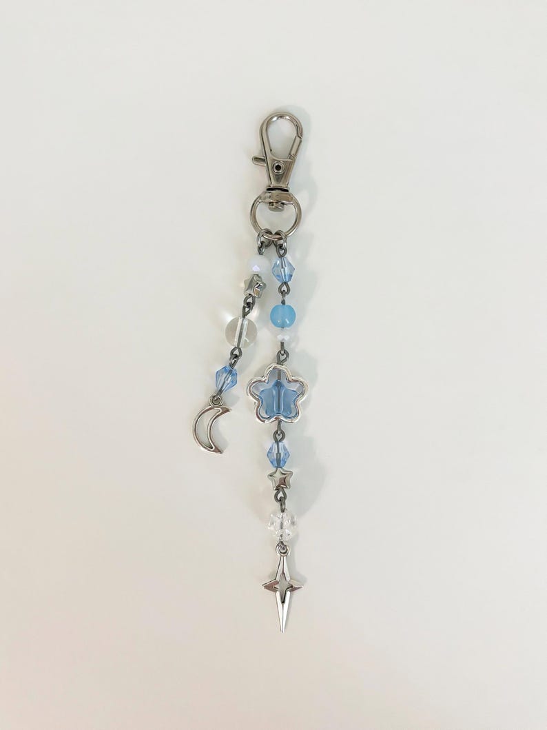 May include: A silver-toned keychain with a lobster clasp. The charm features a crescent moon, a blue flower, a star, and clear and blue beads. The keychain is designed with celestial elements.