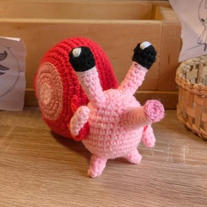 May include: A handmade crochet snail figurine. The snail has a red shell and a pink body with black and white eyes. The snail is standing on a wooden surface.