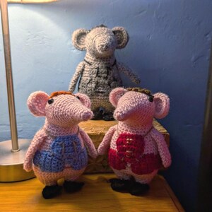 Puede incluir: Tres ratones de ganchillo hechos a mano. Dos ratones rosas visten chalecos, uno azul y otro rojo. Un ratón gris más grande está detrás. Todos tienen pies negros y detalles marrones. Una exhibición artesanal encantadora.