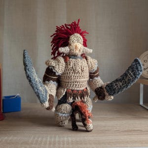 May include: A handmade crochet warrior doll with a red mohawk, tan armor, and two gray swords. The doll has brown and tan accents, and is posed on a wooden surface. The doll is a unique, handcrafted item.