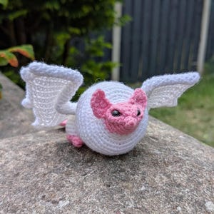 Puede incluir: Un murciélago de ganchillo blanco con detalles en rosa. El murciélago tiene un cuerpo redondo, orejas rosas y pequeños ojos negros. Sus alas están extendidas y está posado sobre una superficie de piedra. Un objeto artesanal y adorable.