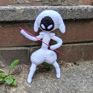 May include: A handmade white crochet doll with a black face and a pink crochet needle. The doll has a unique design with a bulbous body and long legs. The doll is standing on a concrete surface.