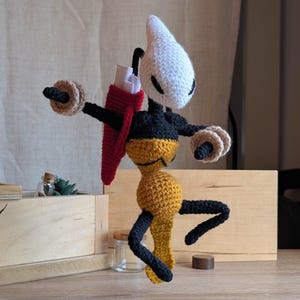 Può includere: Bambola all'uncinetto fatta a mano con testa bianca, corpo nero e parte inferiore gialla. Ha braccia e gambe nere e tiene in mano due oggetti circolari color beige. Una struttura rossa a forma di zaino è attaccata alla schiena.