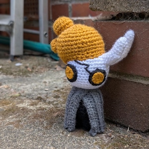 May include: A handmade crochet figure featuring a yellow head with two small pom-poms, a white face with black and yellow eyes, and a gray cloak. The figure is leaning against a brick wall, showcasing its detailed craftsmanship.