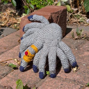 May include: A handmade gray crochet spider with eight legs, each tipped with navy blue. The spider has a yellow and red embellishment on one leg. The spider is resting on a red brick surface.