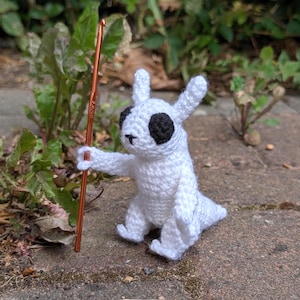 May include: A handmade white crochet creature with black eyes and a small black nose, holding a copper crochet hook. The creature is sitting on a brick surface, with green plants in the background. The crochet hook is approximately 6 inches long.