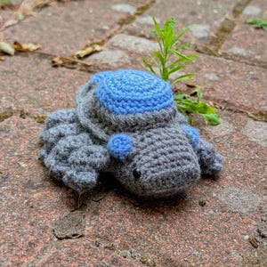 May include: A crocheted turtle toy in grey and blue. The turtle features a grey body, a blue shell, and blue details. The handmade turtle is resting on a brick surface, highlighting the texture of the crochet.