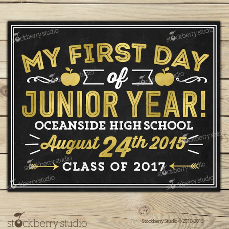 Op de afbeelding: Een schoolbord-stijl bord met gouden letters en accenten. De tekst luidt "MY FIRST DAY of JUNIOR YEAR! OCEANSIDE HIGH SCHOOL August 24th 2015 CLASS OF 2017". Het bord heeft een witte rand en appel illustraties.