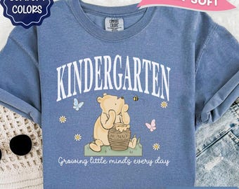 May include: A blue short-sleeve t-shirt featuring a graphic of a yellow bear sitting by a honey pot. The text reads KINDERGARTEN and Growing little minds every day. The design includes small flowers, butterflies, and a bee on the fabric.