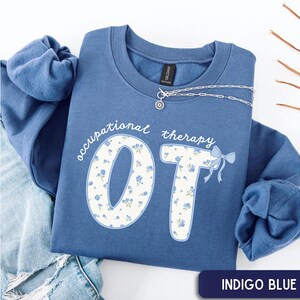 May include: Indigo blue sweatshirt with the words "occupational therapy OT" in white script and floral print. The "OT" letters are filled with a blue floral pattern and a blue bow. A silver necklace is draped on the sweatshirt.