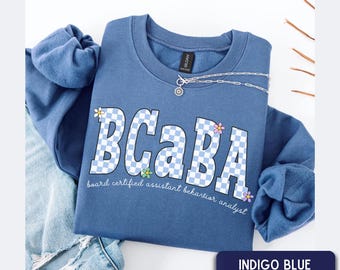 May include: An indigo blue crewneck sweatshirt featuring the text BCABA in a light blue and white checkered pattern. Small colorful flower graphics are placed around the letters. Below the main text, the words board certified assistant behavior analyst are printed in a white script font. A silver chain necklace with a circular pendant rests on the neckline.