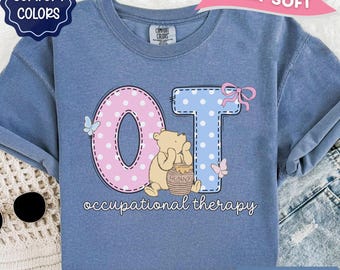 May include: A blue jean colored t-shirt featuring a graphic with the letters OT in pink and blue polka dots. A light brown bear character holds a honey pot labeled Hunny. The text occupational therapy is printed below with butterfly accents.