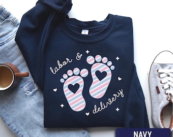 May include: A navy blue crewneck sweatshirt featuring a graphic of two baby footprints with diagonal pink and blue stripes and a heart cutout in the center of each foot. The text "labor & delivery" is written in white cursive script.