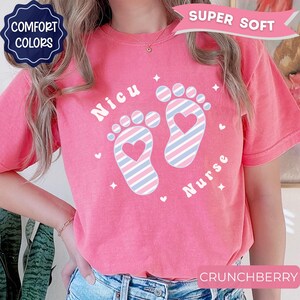 May include: A coral pink short-sleeve t-shirt featuring a graphic of two baby footprints with diagonal blue, pink, and white stripes. The text 'Nicu' and 'Nurse' is printed in white, accompanied by small white heart and star icons.