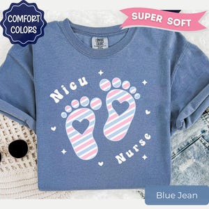 May include: A blue crew neck t-shirt featuring a graphic of two baby footprints with diagonal pink and blue stripes and a heart cutout in the center of each foot. The text "Nicu" and "Nurse" is printed in white around the footprints with small stars.