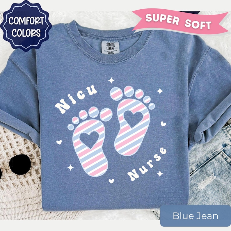May include: A blue crew neck t-shirt featuring a graphic of two baby footprints with diagonal pink and blue stripes and a heart cutout in the center of each foot. The text "Nicu" and "Nurse" is printed in white around the footprints with small stars.