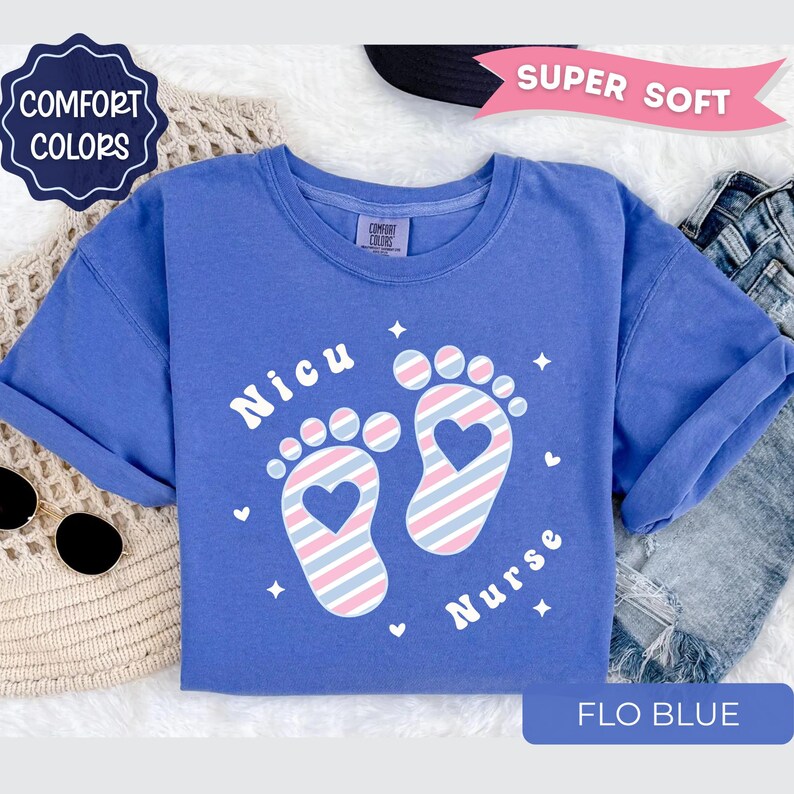 May include: A periwinkle blue t-shirt featuring the text Nicu Nurse and a graphic of two baby footprints with diagonal pink, blue, and white stripes. Each footprint contains a heart cutout, surrounded by small white star and heart icons.