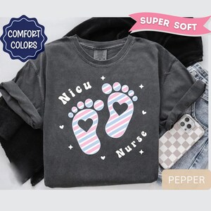 May include: A charcoal gray t-shirt featuring a graphic of two baby footprints with a diagonal pink and blue striped pattern and a heart cutout in each heel. The text "Nicu" and "Nurse" is printed in white, accompanied by small white star and heart icons.