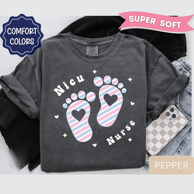 May include: A charcoal gray t-shirt featuring a graphic of two baby footprints with a diagonal pink and blue striped pattern and a heart cutout in each heel. The text "Nicu" and "Nurse" is printed in white, accompanied by small white star and heart icons.