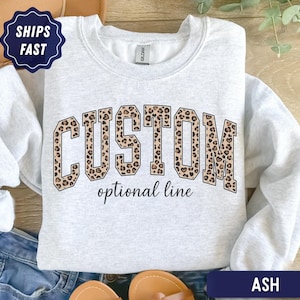 May include: An ash gray crewneck sweatshirt featuring the word CUSTOM in large, block-style letters with a brown and black leopard print pattern. Below the word CUSTOM, the text optional line is printed in a smaller, black cursive font.