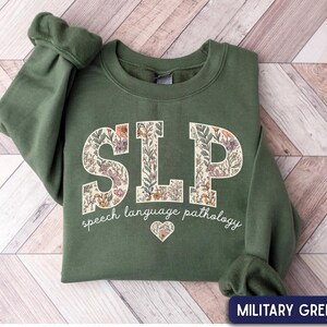May include: A military green crewneck sweatshirt featuring the letters SLP in a large block font filled with a floral pattern. Below the letters, the text reads speech language pathology in white script, followed by a small floral heart icon.