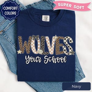 May include: A navy blue t-shirt featuring the text WOLVES in a mix of leopard print, checkered, and polka dot patterns, with a small bow, smiley face, and star accents. Below, the text reads Your School in white cursive script lettering.