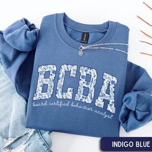 May include: An indigo blue crewneck sweatshirt featuring the letters BCBA in a white floral pattern. Below the letters, the text reads board certified behavior analyst in a white script font. A silver chain necklace rests on the collar.