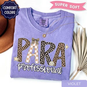 May include: A violet short-sleeve t-shirt with a graphic print that reads "PARA professional" in large letters. The letters feature leopard print, cheetah print, and a tan checkerboard pattern with a small bow and smiley face icon.