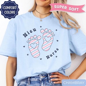 May include: A light blue t-shirt featuring a graphic of two baby footprints with pink and blue diagonal stripes and a heart cutout in the center of each. The text "Nicu" and "Nurse" is printed in dark blue, accompanied by small star and heart icons.