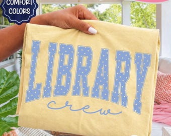 Peut inclure: Un t-shirt jaune pâle avec le texte LIBRARY CREW en grandes lettres majuscules bleu clair à pois blancs. Le mot LIBRARY est imprimé dans une police de style universitaire, tandis que le mot crew est écrit en lettres cursives.
