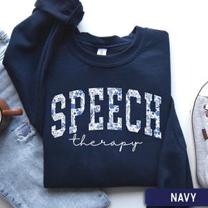 May include: A navy blue crewneck sweatshirt featuring the word SPEECH in large, block-style letters filled with a blue and white floral pattern, with the word therapy written below in a white, cursive script font. The garment is a solid navy color.