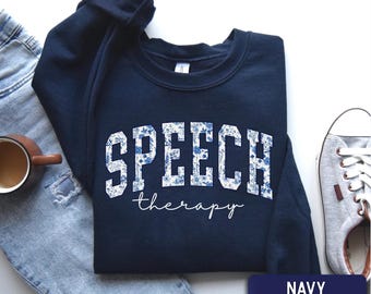 May include: A navy blue crewneck sweatshirt featuring the word SPEECH in large, block-style letters filled with a blue and white floral pattern, with the word therapy written below in a white, cursive script font. The garment is a solid navy color.
