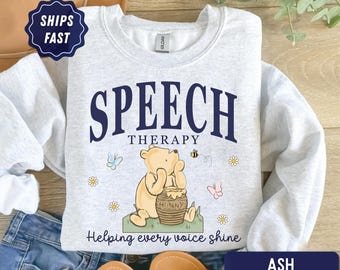 Może przedstawiać: Popielata bluza z okrągłym dekoltem i ciemnoniebieską grafiką z napisem SPEECH THERAPY oraz Helping every voice shine. Wzór przedstawia żółtego misia siedzącego przy garnku z miodem, otoczonego małymi kwiatkami, motylami i pszczołą.
