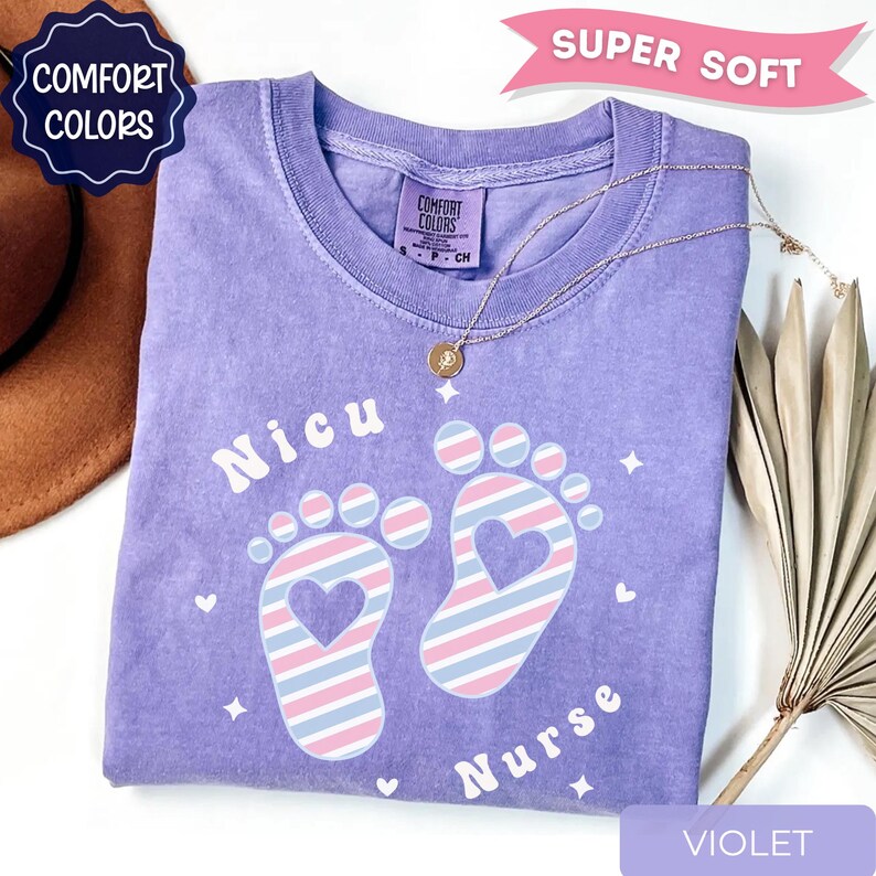 May include: A violet short-sleeved t-shirt with a graphic print featuring two baby footprints filled with diagonal pink, blue, and white stripes and heart-shaped cutouts. The text reads Nicu Nurse, accompanied by small star and heart icons.