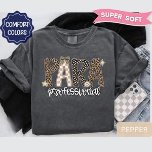 May include: A charcoal gray t-shirt featuring the text PARA professional in a mix of leopard print, cheetah print, and tan checkerboard patterns. The design includes a small bow, a smiley face, and star accents. A pink banner reads SUPER SOFT.