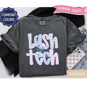 May include: A dark gray t-shirt with the words "LASH TECH" in a floral and gingham pattern. The shirt has a crew neck and long sleeves. A phone with a checkered case is visible.