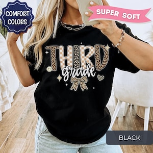 May include: A black t-shirt featuring the text THIRD grade in a mix of tan checkered and leopard print patterns. The design includes a leopard print bow, small stars, a heart, and a smiley face. Text labels include COMFORT COLORS and SUPER SOFT.