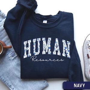 May include: A navy blue crewneck sweatshirt featuring the word HUMAN in large block letters filled with a blue and white floral pattern, with the word Resources written below in a white script font. The sweatshirt is displayed with jeans and a sneaker.
