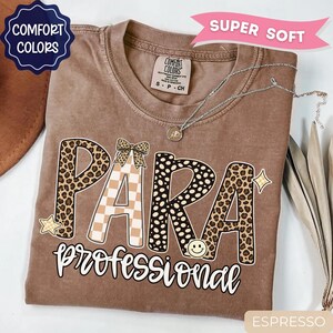 May include: A brown Comfort Colors t-shirt featuring the text PARA in large letters with leopard print and checkered patterns, a small bow, and a smiley face. Below, the word professional is written in white script. Includes a silver necklace.