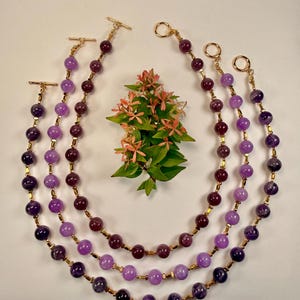May include: Three layered necklaces with amethyst beads in varying shades of purple and gold-colored accents. The necklaces are arranged in a semi-circle, with a small floral arrangement in the center.