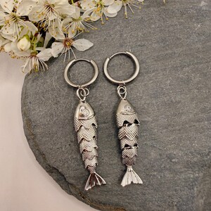 Può includere: Un paio di orecchini argentati a forma di pesce con chiusura a cerchio. Gli orecchini presentano un design a scaglie dettagliato e una piccola gemma sulla testa. Gli orecchini sono esposti su una superficie di pietra grigia, con fiori bianchi sullo sfondo.