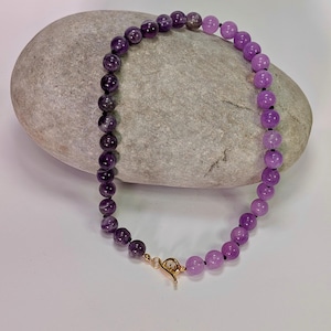 Puede incluir: Un collar de cuentas con un degradado de tonos morados. El collar presenta cuentas redondas que transicionan de un morado oscuro y profundo a un tono lavanda más claro. Un cierre dorado asegura el collar. El collar se muestra sobre una piedra gris.