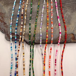 May include: Several beaded necklaces in a range of colours, such as blue, green, orange, and red. The necklaces are made with small, colourful beads and are shown against a dark, textured backdrop. The beads are strung together to create a delicate, linear design.