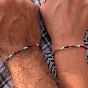 Könnte beinhalten: Zwei Armbänder mit Perlen, jedes mit einem Muster aus roten, weißen, grünen und schwarzen Perlen. Die Armbänder werden an Handgelenken getragen, eines an einer Person mit behaarten Armen und das andere an einer Person mit glatter Haut.