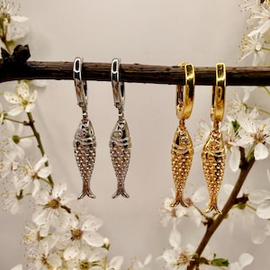 Op de afbeelding: Twee paar visvormige oorbellen, één zilver en één goud, hangen aan een bruine tak. De oorbellen hebben een gedetailleerd schubbenpatroon en zijn bevestigd aan oorringsluitingen. Witte bloemen met gele centra zijn op de achtergrond.